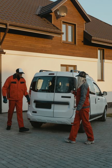 A man wearing a black cap, face mask, beige and gray jacket, gray pants, and sneakers stands next to a white commercial van parked on a narrow brick-paved residential street in Clapton, E5. The van is
