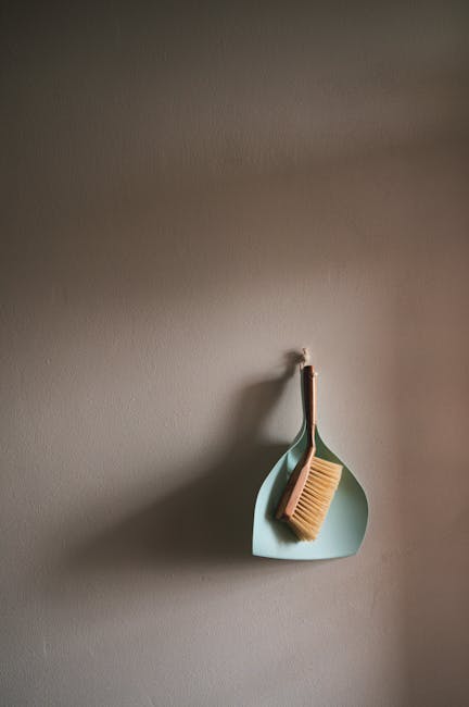 A minimalist cleaning tool setup on a neutral-toned wall featuring a pale green dustpan and a wooden-handled scrub brush with natural bristles, hanging from a small hook. The dustpan is slightly curve