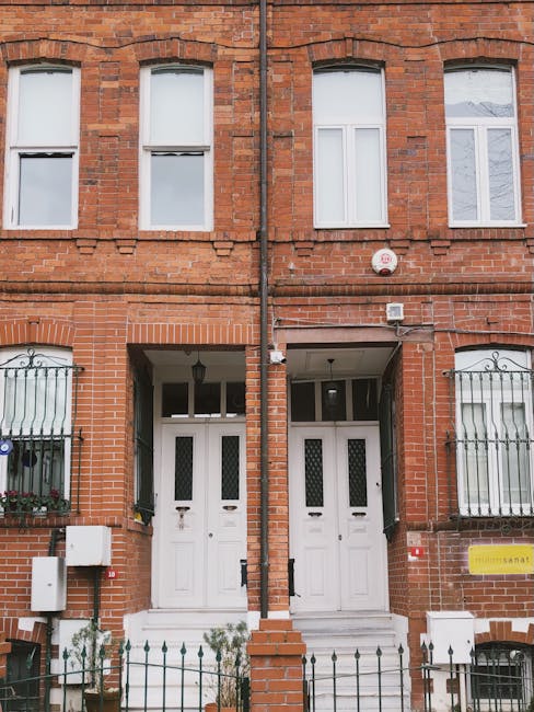 The image depicts the exterior of a red brick residential building with two white front doors, each with a small window near the top. The building has three upper-floor windows with white frames, evenly spaced, and the windows are clean and free of dirt or grime. Decorative black iron bars are installed on some windows, and a small white security alarm box is mounted on the wall. The ground level features small front gardens enclosed by black metal railings, with some greenery visible near the entrances. The environment is well-maintained and clean, reflecting good hygiene and upkeep typical of property management. This setting illustrates the kind of façade that professional cleaning services like Carpet Cleaners E5 can help maintain through regular or deep cleaning, ensuring surfaces remain spotless and presentable for tenants or visitors.