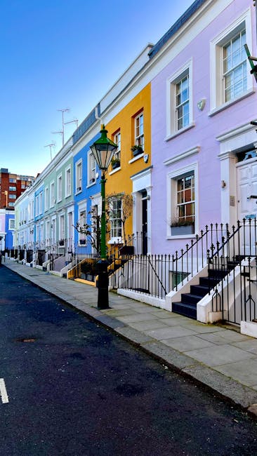 A row of pastel-coloured terraced houses on a quiet street in Mare Street Clapton Common E5, featuring facades painted in shades of blue, yellow, and lavender. The street has a smooth asphalt road with a concrete sidewalk. Classic black wrought-iron railings decorate the front steps of each home, leading to front doors with white frames and small windows. A traditional street lamp with a green and black finish stands near the pavement, and some houses have window boxes with plants. Bright daylight illuminates the scene, highlighting the clean and well-maintained exterior surfaces of the buildings, reflecting a tidy, residential environment. Carpet Cleaners E5 provides expert domestic cleaning and surface sanitisation services, ensuring high standards of hygiene and cleanliness for homes like these.