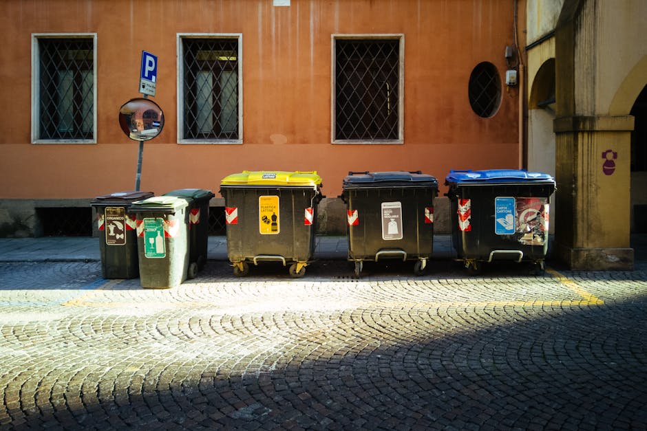 A pile of overflowing waste bins and scattered rubbish on a sidewalk in front of a commercial building with a blue metal staircase and storefronts, including a fish bar, in the background. The waste i