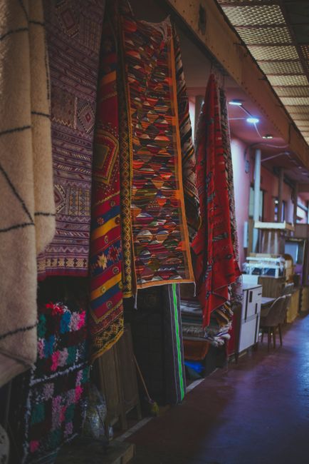 An interior view of a rug showroom showcasing a variety of colorful, patterned carpets and rugs hanging vertically along a wall. The rugs feature intricate geometric and floral designs in vibrant reds, blues, and other rich hues. The space is illuminated with warm lighting, highlighting the textures and details of the textiles. Below the hanging carpets, additional rolled or folded rugs are stacked on a display stand. The room has a wooden floor and shelves with other home decor items, suggesting a retail environment. Carpet Cleaners E5 provides surface cleaning and deep cleaning services for such textiles, ensuring optimal hygiene and preservation, as seen in this scene from their Hackney Wick location at Broadway Market, E5.