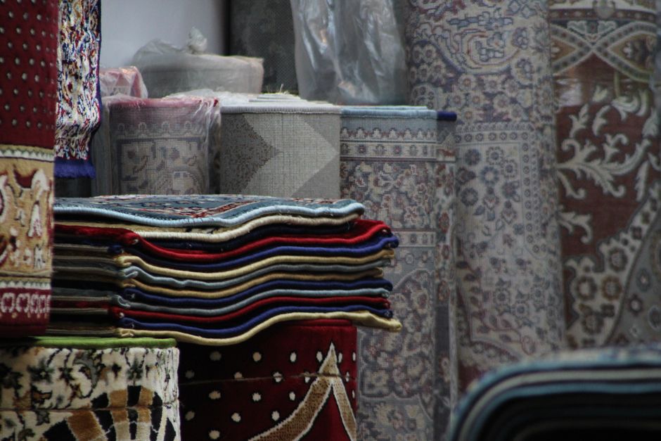 An interior view of a rug showroom showcasing a variety of colorful, patterned carpets and rugs hanging vertically along a wall. The rugs feature intricate geometric and floral designs in vibrant reds, blues, and other rich hues. The space is illuminated with warm lighting, highlighting the textures and details of the textiles. Below the hanging carpets, additional rolled or folded rugs are stacked on a display stand. The room has a wooden floor and shelves with other home decor items, suggesting a retail environment. Carpet Cleaners E5 provides surface cleaning and deep cleaning services for such textiles, ensuring optimal hygiene and preservation, as seen in this scene from their Hackney Wick location at Broadway Market, E5.