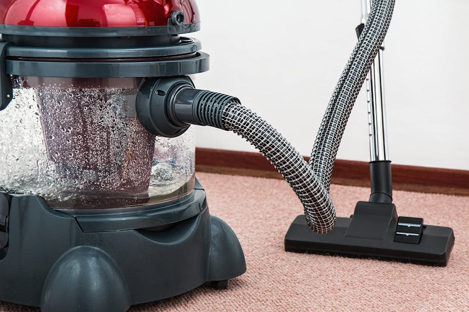 A woman using a yellow vacuum cleaner with a black hose to deep clean a patterned area rug in a living room. The room features a wooden floor, a red leather sofa, and natural lighting. The woman is dr