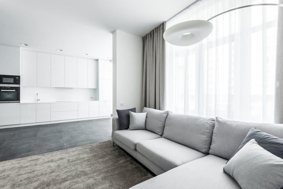 A modern, open-plan living room with a light grey fabric sofa adorned with matching cushions, positioned near large, sheer white curtains that allow natural light to fill the space. The room features a neutral-toned carpet on the floor, contrasting with the sleek, dark grey tiled kitchen area in the background, equipped with built-in white cabinets, stainless steel appliances, and a clean, minimalist design. The overall ambiance is bright and tidy, emphasizing cleanliness and tidy maintenance, consistent with professional domestic cleaning standards. Natural daylight highlights the polished surfaces and pristine condition of the upholstery, reflecting thorough surface cleaning and deep sanitation provided by Carpet Cleaners E5 near Hackney Central station.